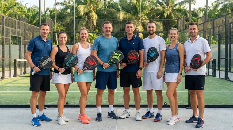 Группа Padel Camp на корте: девять человек с ракетками стоят в ряд, за ними пальмы и стеклянная стена корта